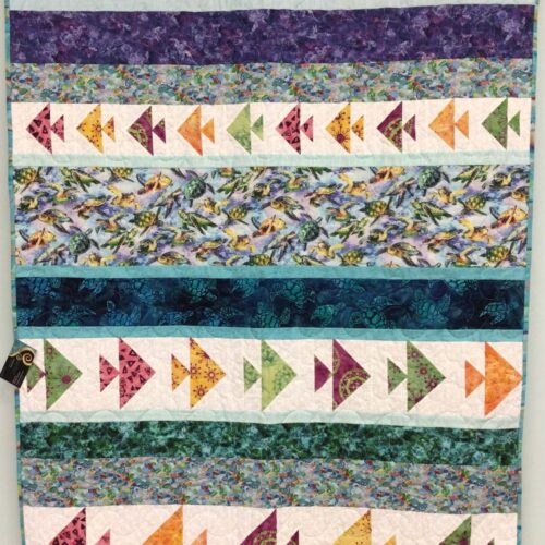 Lap quilt with horizontal stripes alternating between ocean-themed prints and pieced fish blocks in various colors with sea turtles and tropical fish
