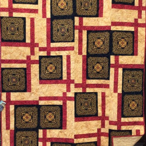 Machine-pieced throw quilt featuring gold filigree squares with red and gold accents and a gold feathery leaf print backing.