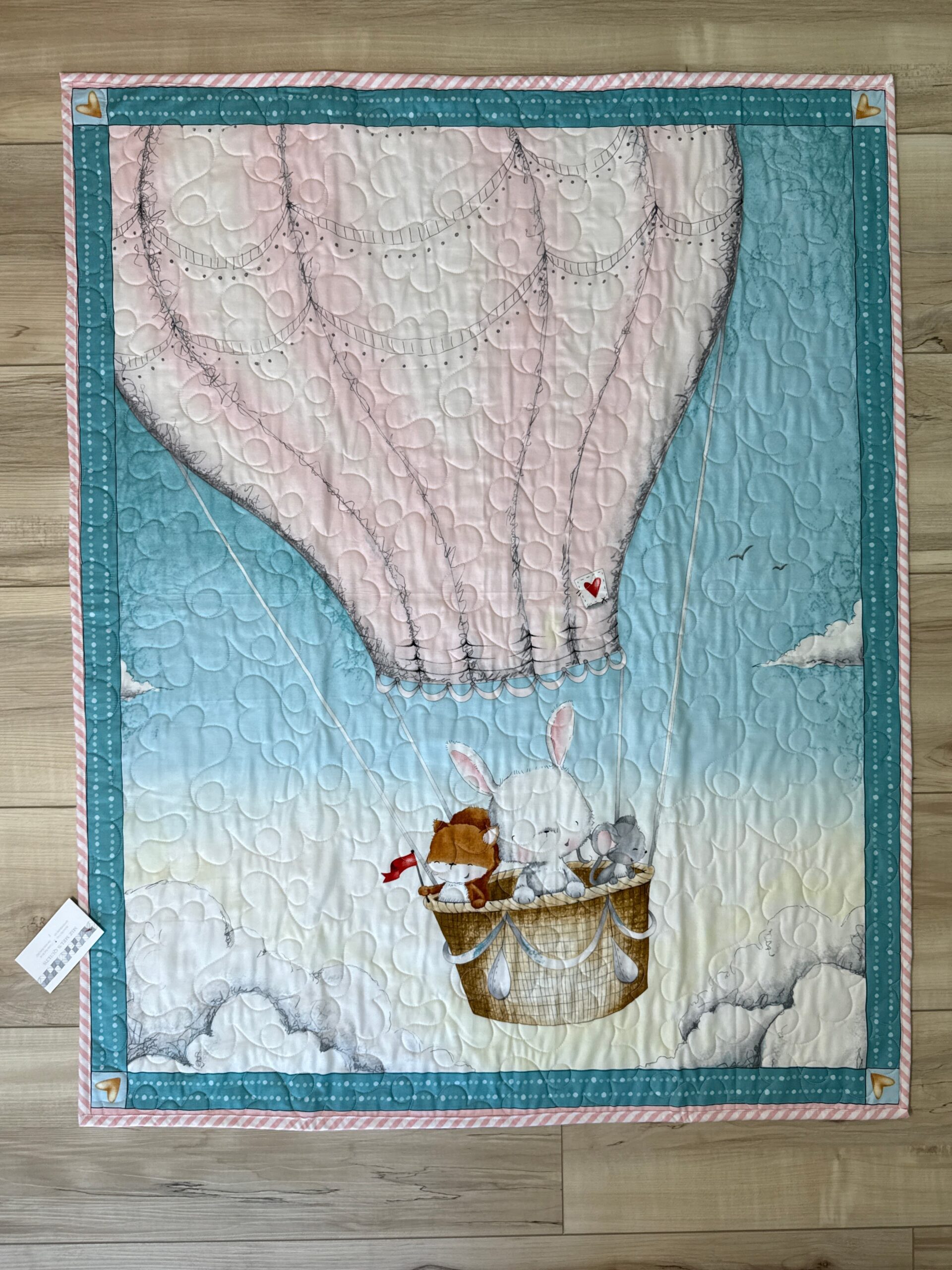 Baby quilt featuring bunny and friends in cream hot air balloon on soft blue sky background with teal borders and long arm quilting