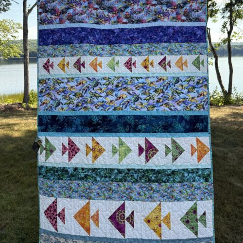 Lap quilt with horizontal stripes alternating between ocean-themed prints and pieced fish blocks in various colors with sea turtles and tropical fish