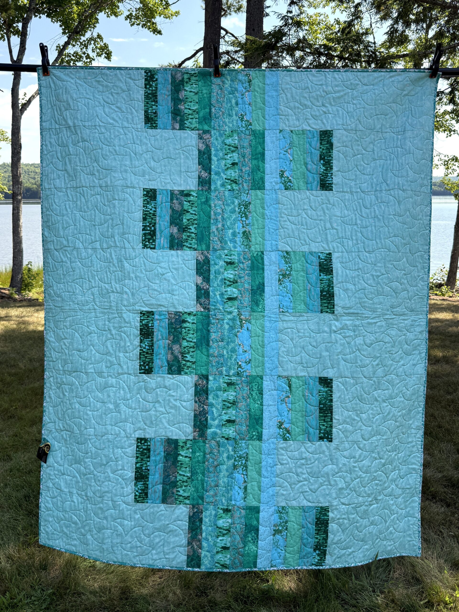 Modern throw quilt featuring vertical strips of varied teal fabrics against light teal background with all over long arm quilting