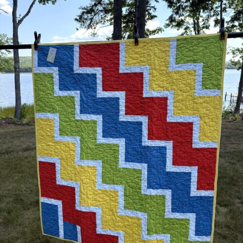 Lap quilt with interlocking zigzag blocks in primary colors red, yellow, blue, and green with white sashing and polka dot backing