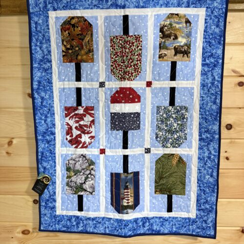 Wall hanging with nine buoy-shaped blocks featuring Maine motifs including moose, lobsters, lighthouse, and wild blueberries