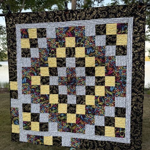 Throw quilt with Trip Around the World pattern featuring small squares in music-themed fabrics radiating from center in concentric rings