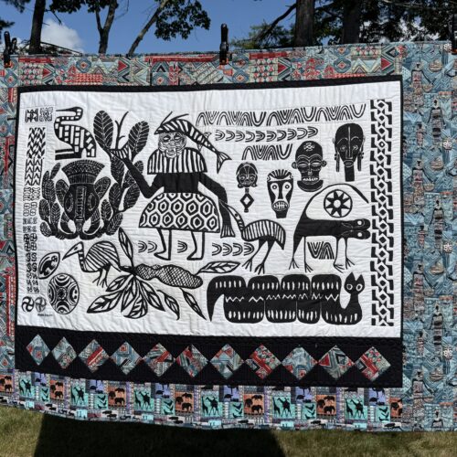 Wall hanging featuring African Ashia batik panel in black and white with tribal masks, animals, and geometric designs bordered with pieced blocks