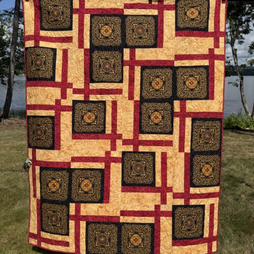 Machine-pieced throw quilt featuring gold filigree squares with red and gold accents and a gold feathery leaf print backing.