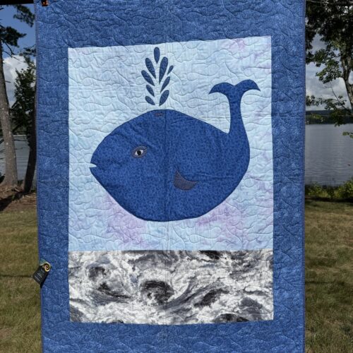 Baby quilt featuring friendly navy whale with water spout appliqué on watercolor background with marbled ocean waves and blue purple gingham backing