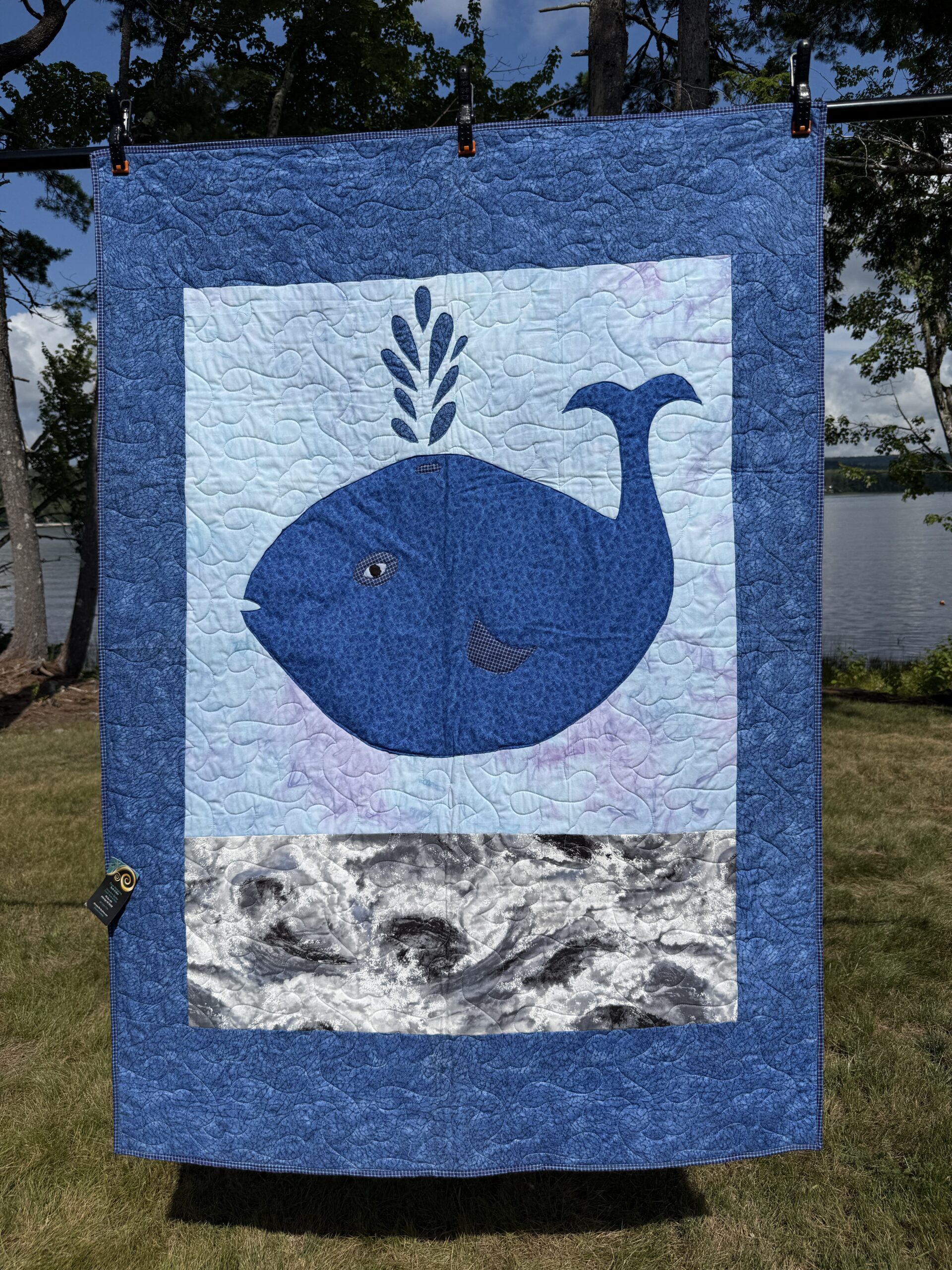 Baby quilt featuring friendly navy whale with water spout appliqué on watercolor background with marbled ocean waves and blue purple gingham backing