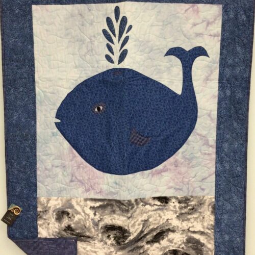 Baby quilt featuring friendly navy whale with water spout appliqué on watercolor background with marbled ocean waves and blue purple gingham backing