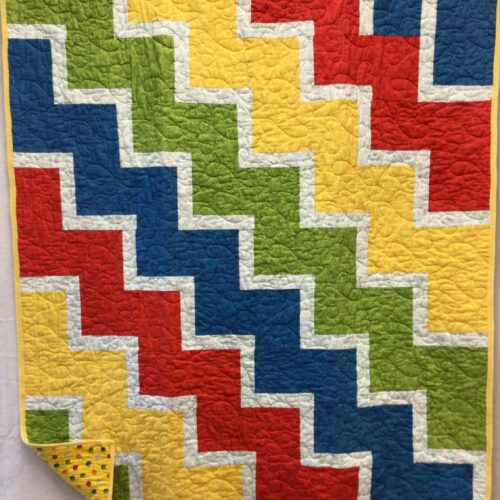 Lap quilt with interlocking zigzag blocks in primary colors red, yellow, blue, and green with white sashing and polka dot backing