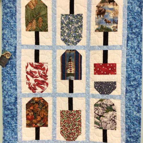 Wall hanging with nine buoy-shaped blocks featuring Maine motifs including moose, lobsters, lighthouse, and wild blueberries