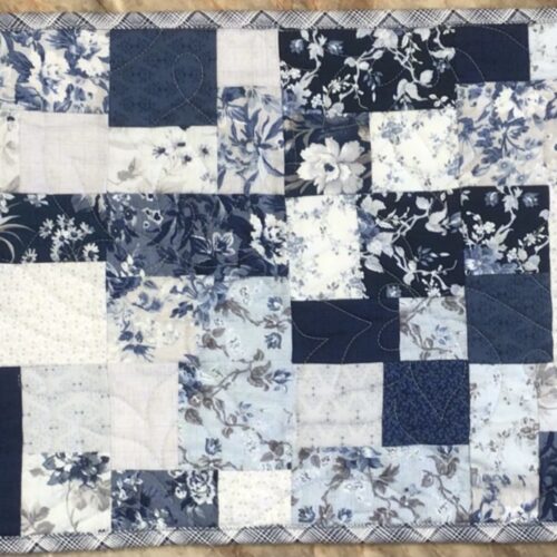 Handmade quilted table runner in blue and white patchwork featuring assorted floral prints, finished with longarm quilting in the Leaf Crown pattern.