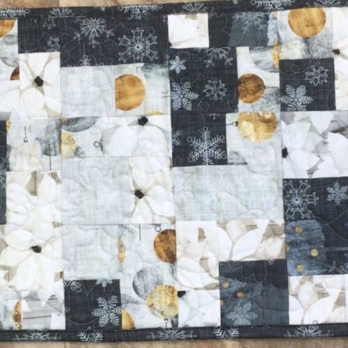 Handmade quilted table runner in charcoal gray, white, and gold patchwork featuring snowflake and white floral prints, finished with longarm quilting in a loops and swirls pattern.
