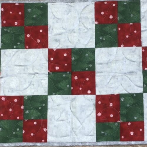 Handmade quilted Christmas table runner with red, green, and silver-gray patchwork squares, each featuring small scattered snowflake prints.