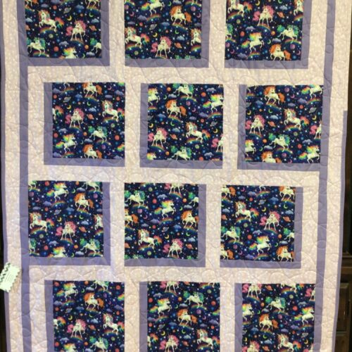 Handmade baby quilt with twelve unicorn print blocks featuring unicorns, rainbows, planets, and stars on a navy background, framed in soft pink and purple fabrics.