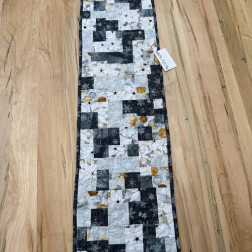 Handmade quilted table runner in charcoal gray, white, and gold patchwork featuring snowflake and white floral prints, finished with longarm quilting in a loops and swirls pattern.