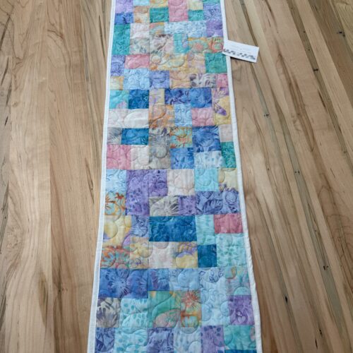 Handmade quilted table runner in soft pastel batik patchwork featuring lavender, peach, mint, and light blue tones with a circular quilting motif.