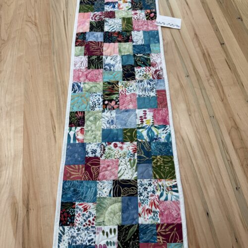 Handmade quilted table runner in vibrant multicolor batik patchwork featuring tropical leaf and floral prints in teal, pink, green, and coral tones.