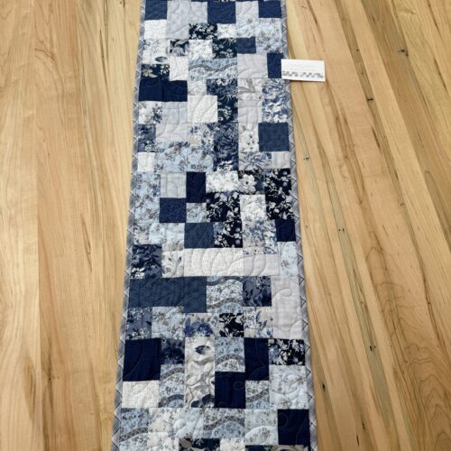 Handmade quilted table runner in blue and white patchwork featuring assorted floral prints, finished with longarm quilting in the Leaf Crown pattern.