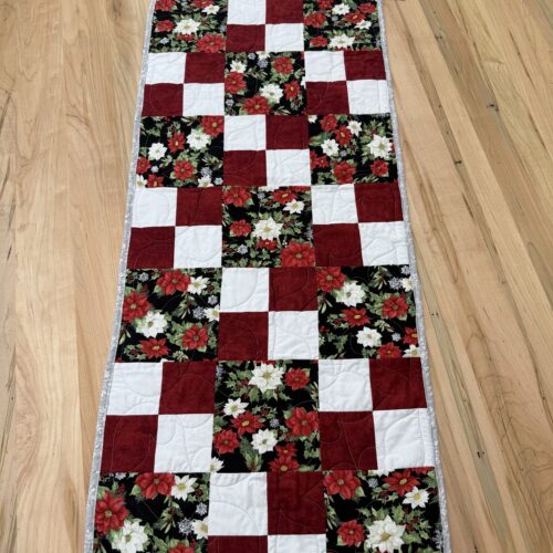 Handmade quilted Christmas table runner with red and white poinsettia print blocks alternating with solid burgundy and white squares, finished with silver-gray binding.
