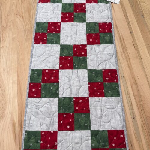 Handmade quilted Christmas table runner with red, green, and silver-gray patchwork squares, each featuring small scattered snowflake prints.