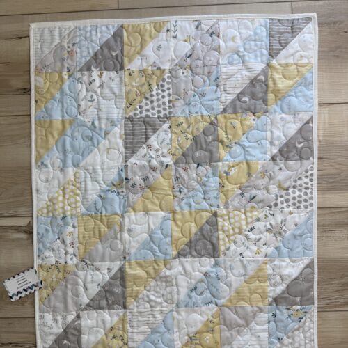 Handmade baby quilt with half-square triangle patchwork in soft yellow, light blue, gray, and white nursery prints featuring moons, clouds, and botanical motifs.