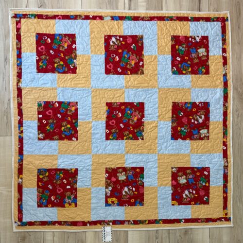 Handmade baby quilt with nine red teddy bear print blocks framed in golden yellow and light blue fabric, pieced in the Town Square pattern by Fabric Cafe.