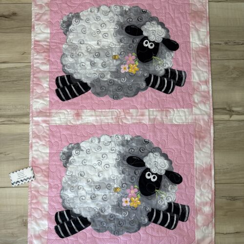 Handmade pink baby quilt featuring two large panel-printed cartoon sheep with curly fleece, holding flowers, on a pink polka dot background.