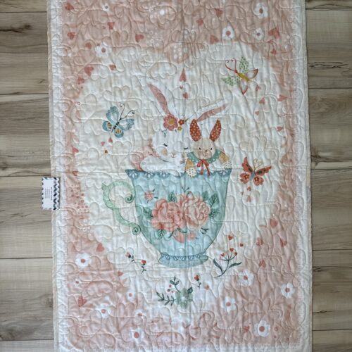 Handmade panel baby quilt with two bunnies sitting in a teal rose-decorated teacup, surrounded by butterflies on a soft pink floral background.