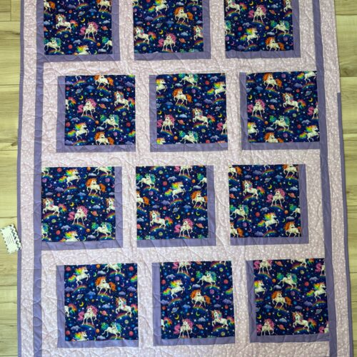 Handmade baby quilt with twelve unicorn print blocks featuring unicorns, rainbows, planets, and stars on a navy background, framed in soft pink and purple fabrics.