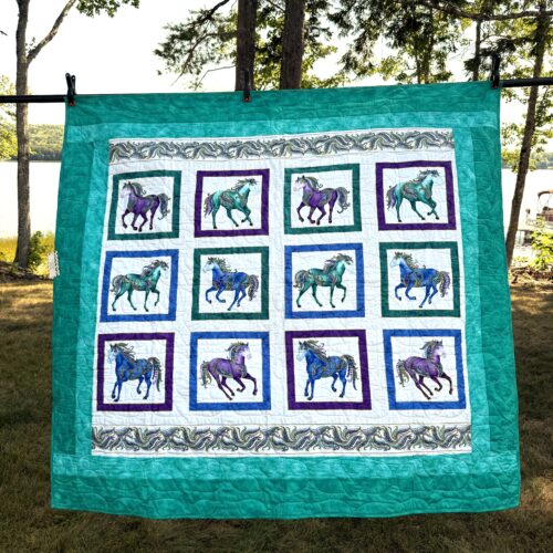 Handmade lap quilt with twelve decorative carousel horses in teal, blue, and purple, framed individually and set against multiple teal borders.