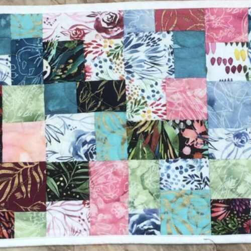 Handmade quilted table runner in vibrant multicolor batik patchwork featuring tropical leaf and floral prints in teal, pink, green, and coral tones.