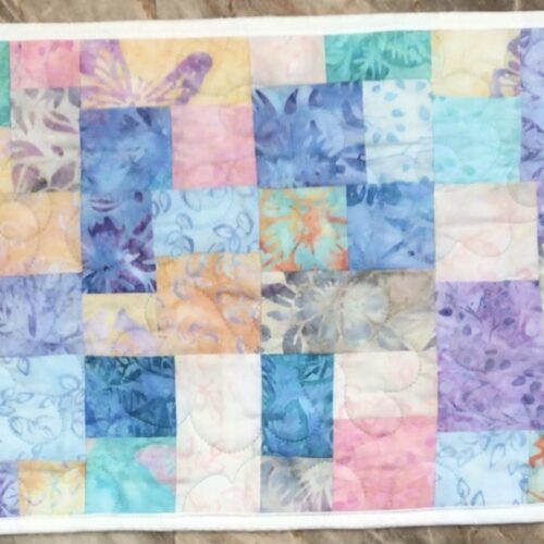 Handmade quilted table runner in soft pastel batik patchwork featuring lavender, peach, mint, and light blue tones with a circular quilting motif.