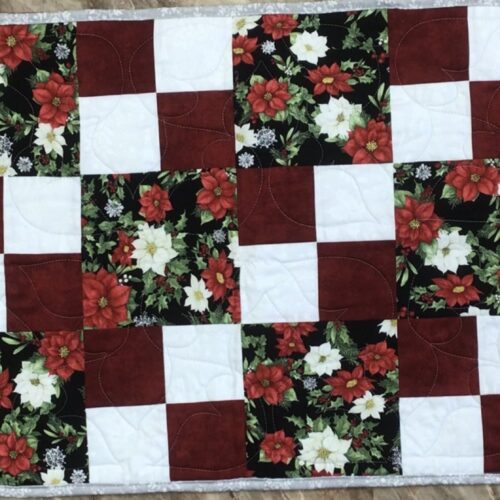Handmade quilted Christmas table runner with red and white poinsettia print blocks alternating with solid burgundy and white squares, finished with silver-gray binding.