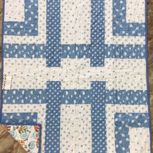 Handmade baby quilt in blue and white woven strip piecing with small heart, kite, and cloud prints, backed with a colorful sailboat print.