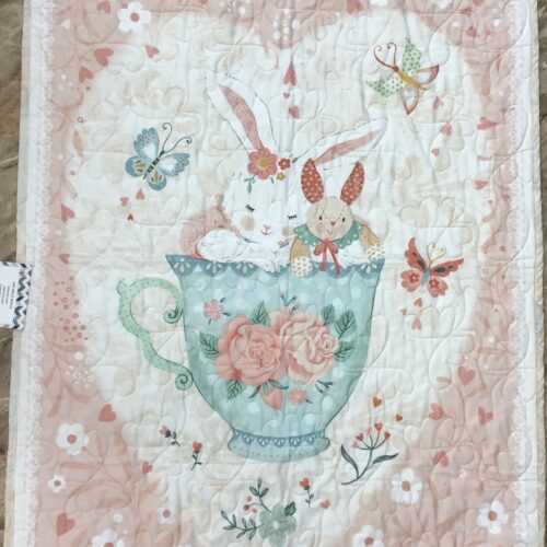 Handmade panel baby quilt with two bunnies sitting in a teal rose-decorated teacup, surrounded by butterflies on a soft pink floral background.